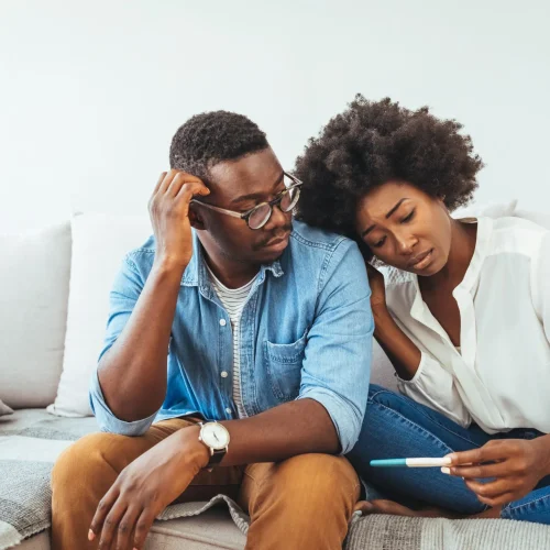 A couple sits together on a couch, looking concerned while the woman holds a pregnancy test. The man touches his head with worry, and the woman leans on his shoulder, appearing anxious and thoughtful.