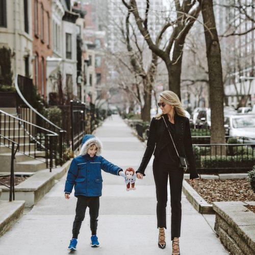 A woman in black attire and sunglasses walks on a city sidewalk, holding hands with a young boy in a blue coat and hood. The boy is holding a small stuffed toy. Leafless trees line the street on an overcast day.