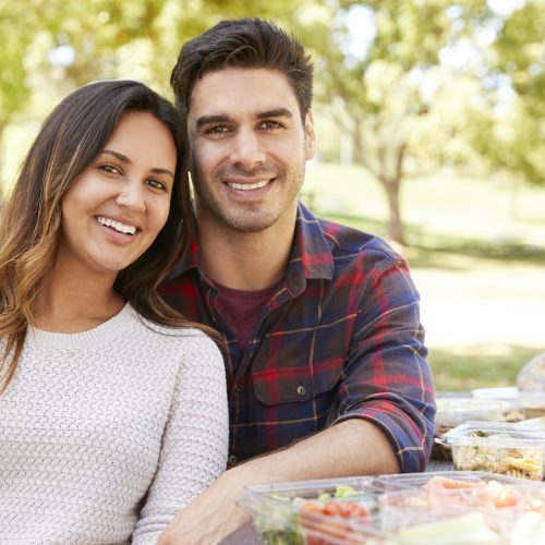 A smiling couple sits close together at a picnic table outdoors, surrounded by various food containers, with trees and greenery in the background.