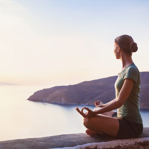 A woman sits cross-legged in a yoga pose on a ledge, facing a calm sea during sunset, with hills in the background and soft sunlight illuminating the scene.