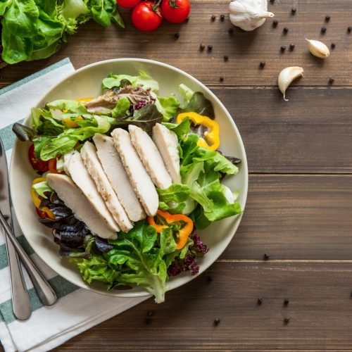 A plate of fresh salad with sliced grilled chicken breast, mixed greens, yellow bell pepper, and cherry tomatoes on a wooden table, with utensils and a napkin beside it. Garlic, tomatoes, and peppercorns are scattered nearby.