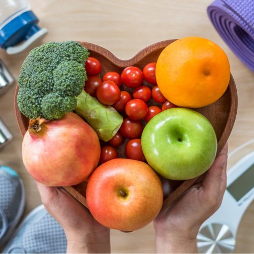 Hands hold a heart-shaped bowl filled with fresh fruits and vegetables, surrounded by fitness items including a yoga mat, dumbbells, sneakers, a water bottle, and a bathroom scale.