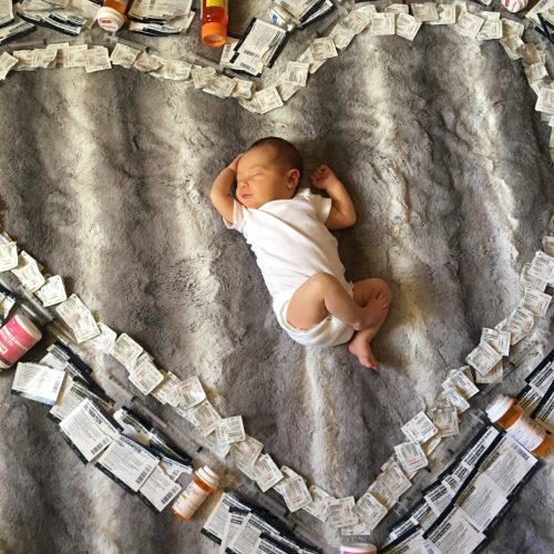 A newborn baby in a white onesie lies on a gray blanket, surrounded by a large heart shape made from medical supplies, including pill bottles, medication packets, and syringes.