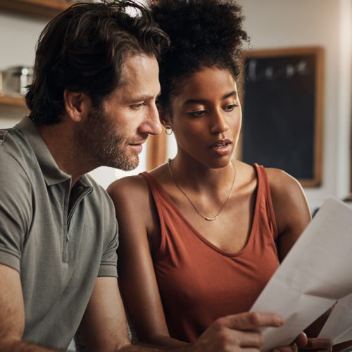 A man and a woman sit together at a kitchen table, looking at documents and a laptop screen with serious expressions. Sunlight comes through the window behind them, and shelves with dishes are visible in the background.