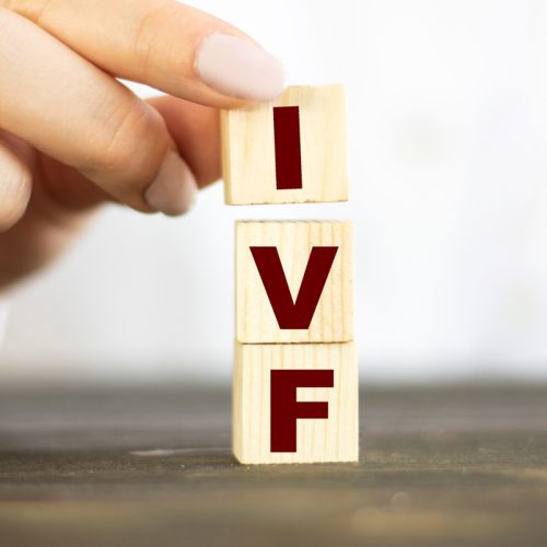 A hand is stacking three wooden blocks with the red letters I, V, and F, forming the abbreviation IVF on a dark surface with a blurred white background.