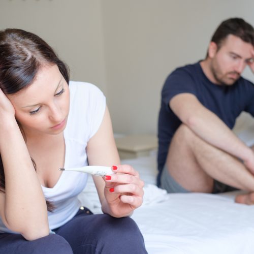 A woman sits on a bed holding a thermometer, looking worried, while a man sits behind her, appearing concerned and pensive. Both have serious expressions in a bright, neutral-colored room.