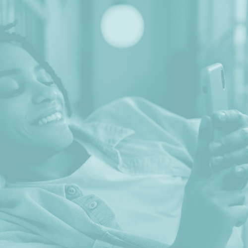 A person smiling and lying on a bed, holding and looking at a smartphone—perhaps checking their end of year fertility insurance options. The image features a teal color overlay.
