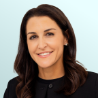 A woman with long brown hair and a black sleeveless top smiles at the camera against a light blue background. She is wearing gold jewelry, including a necklace and earrings.