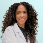A woman with long, curly hair wears a white lab coat and a black shirt, smiling at the camera against a light blue background.
