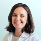 A woman with shoulder-length brown hair smiles at the camera, wearing a white lab coat and a light-colored top, standing against a plain light blue background.