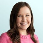 A woman with long brown hair, wearing a bright pink sweater, smiles at the camera against a light blue background.