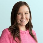 A woman with long brown hair, wearing a bright pink sweater, smiles at the camera against a light blue background.