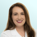 A woman with long brown hair, wearing a white coat and a necklace, smiles at the camera against a light blue background.
