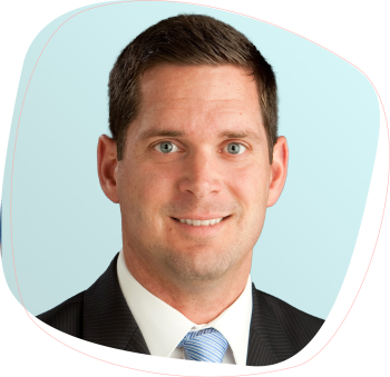 Josh_Swain-Desktop A man with short brown hair, blue eyes, and light skin is smiling. He is wearing a dark suit, white shirt, and light blue striped tie, posed against a light blue background.