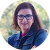 Judith-Becerra A woman with long brown hair and glasses, wearing a denim jacket, stands outdoors with arms crossed and a blurred background of green trees.