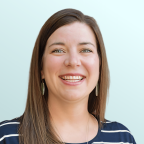 A woman with straight brown hair and green eyes is smiling. She is wearing green earrings and a navy blue top with white stripes, against a light blue background.