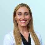 A woman with long, straight blonde hair and blue eyes smiles at the camera. She is wearing a black top and is posed against a plain, light blue background.