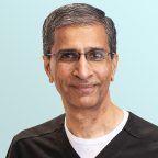 A middle-aged man with short gray hair and glasses, wearing a black shirt over a white undershirt, smiles slightly at the camera against a light blue background.