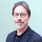 A man with short brown hair, glasses, and a gray goatee is wearing a dark suit and smiling slightly at the camera against a light blue background.