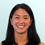 A woman with straight black hair and a floral necklace, wearing a sleeveless black top, smiles in front of a light blue background.