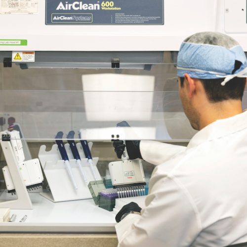 A lab technician in protective clothing works at a biosafety cabinet, using pipettes and handling samples with lab equipment in a sterile environment.