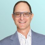 A smiling man wearing clear glasses, a light blue checkered blazer, and a white shirt with blue stripes, posed in front of a light blue gradient background.