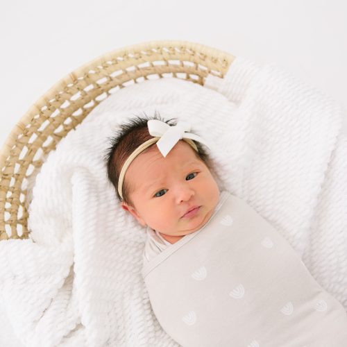 A newborn baby wrapped in a light-colored blanket lies in a woven basket, resting on a textured white blanket, wearing a white headband with a bow.