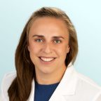 A woman with light brown hair and blue eyes, wearing a white lab coat over a blue top, smiles at the camera in front of a light blue background.