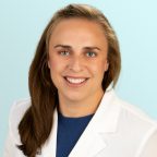 A woman with light brown hair and blue eyes, wearing a white lab coat over a blue top, smiles at the camera in front of a light blue background.