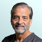 A mature man with gray hair and beard, wearing a navy suit, white shirt, and patterned tie, poses against a light blue background.