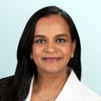 A woman with long dark hair, wearing a white coat, earrings, and a necklace, smiles at the camera against a light blue background.