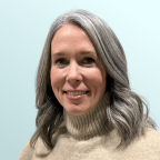 A woman with long, wavy gray hair wearing a beige turtleneck sweater smiles at the camera. The background is a plain, light blue color.