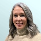 A woman with long, wavy gray hair wearing a beige turtleneck sweater smiles at the camera. The background is a plain, light blue color.