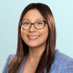 A woman with long brown hair and glasses, wearing a blue blazer and gold necklace, smiles at the camera against a light blue background.