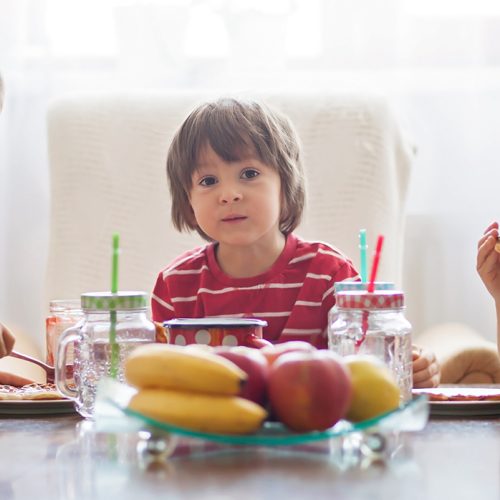 Three young children sit at a table eating together. They all wear red striped shirts. There are plates of food, mason jar drinks with straws, and a fruit bowl with bananas and apples on the table.
