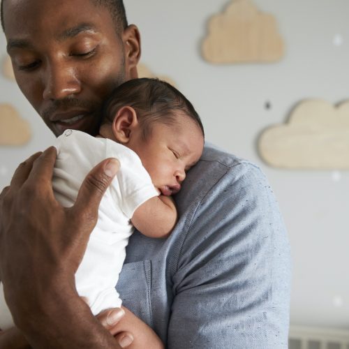 A man gently holds a sleeping newborn baby against his chest in a softly lit nursery with wooden cloud decorations on the wall in the background.