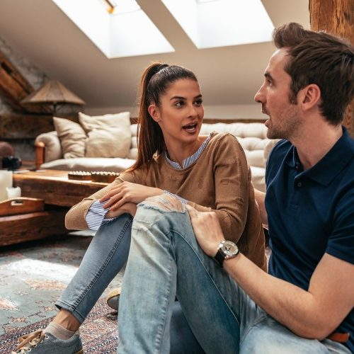A young woman and man sit on the floor in a cozy, rustic living room, engaged in a lively conversation. The room has wooden beams, skylights, and casual decor with a tray and cup on the coffee table.