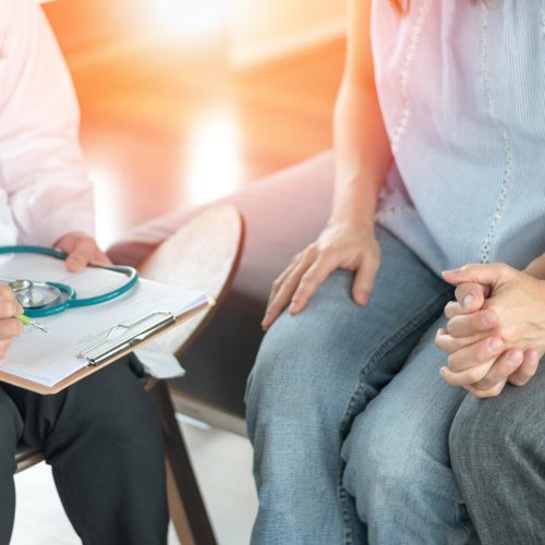 A doctor holding a clipboard and pen speaks with a couple sitting closely together, holding hands, suggesting a medical consultation or discussion about health.