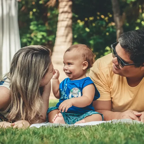 A smiling child sits between two adults on a blanket outdoors, all laughing and enjoying time together on the grass with trees in the background.