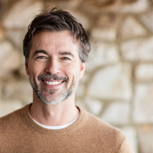 A middle-aged man with short dark hair and a salt-and-pepper beard smiles warmly at the camera. He is wearing a brown sweater and standing in front of a blurred stone wall background.