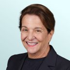 A middle-aged woman with long, light brown hair smiles warmly at the camera. She is wearing a sleeveless black top with beige trim, set against a light blue background.