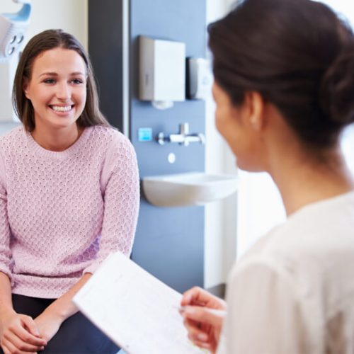 A young woman in a pink sweater smiles while sitting on an exam table, speaking with a healthcare professional holding a clipboard in a medical office.