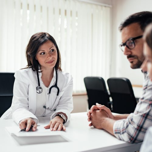 A female doctor in a white coat with a stethoscope talks to a man and a woman sitting across from her at a desk in a medical office with large windows and blinds.