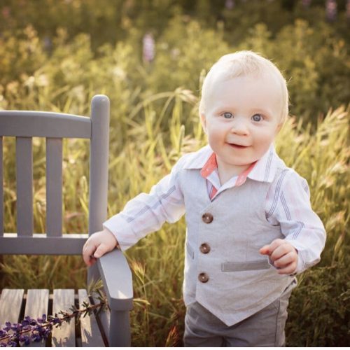 A smiling toddler with blond hair stands next to a gray wooden chair in a sunlit field, wearing a light gray vest, striped shirt, and pink collar. The background is filled with tall green grass and wildflowers.