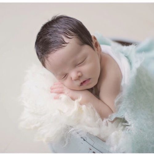 A newborn baby with dark hair sleeps peacefully on a soft white blanket, partially covered with a light blue blanket. The baby is resting on their side with their hands under their cheek.