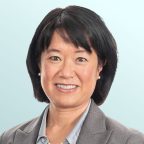 Portrait of an Asian woman with short black hair, wearing a gray blazer, light blue shirt, pearl necklace, and small pearl earrings, smiling in front of a light blue background.