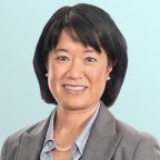 Portrait of an Asian woman with short black hair, wearing a gray blazer, light blue shirt, pearl necklace, and small pearl earrings, smiling in front of a light blue background.