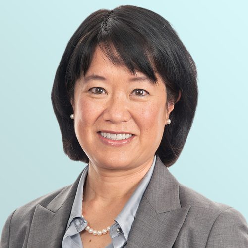 Portrait of an Asian woman with short black hair, wearing a gray blazer, light blue shirt, pearl necklace, and small pearl earrings, smiling in front of a light blue background.