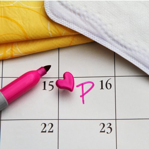 A calendar with the 15th marked by a pink heart pin and the letter P written in pink marker, next to a pink marker, a sanitary pad, and yellow pad packaging.