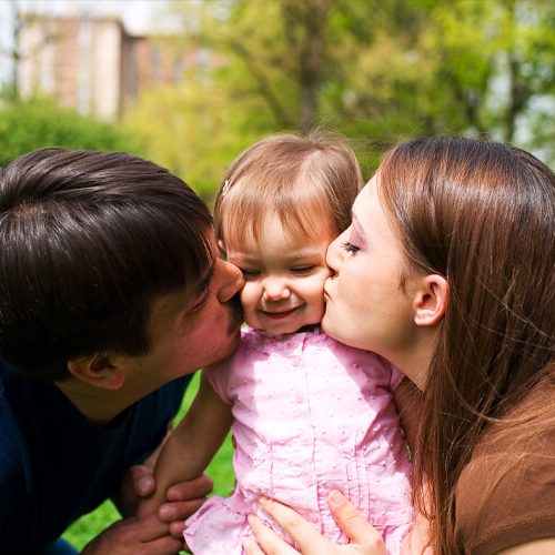 A mother and father kiss their smiling baby daughter on the cheeks while sitting on grass in a park, with trees and a building in the background.